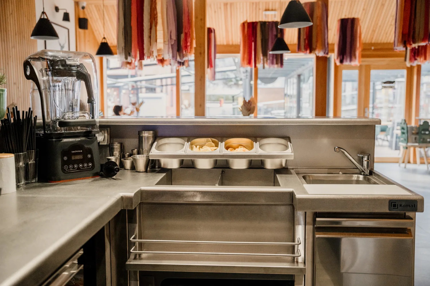 Stanowisko barowe ze stali nierdzewnej z blenderem, pojemnikami na dodatki i zlewem, zaaranżowane w nowoczesnej restauracji.