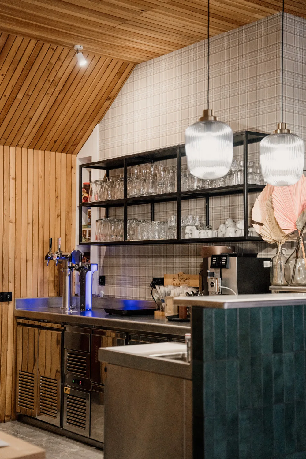 Nowoczesny bar restauracyjny z drewnianą zabudową, wiszącymi lampami, półkami na szkło oraz profesjonalnym zapleczem barowym ze stali nierdzewnej.