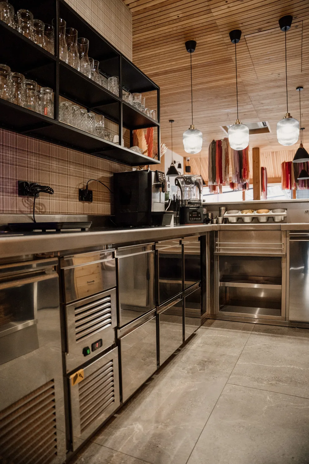 Nowoczesny bar restauracyjny z drewnianą zabudową, wiszącymi lampami, półkami na szkło oraz profesjonalnym zapleczem barowym ze stali nierdzewnej.