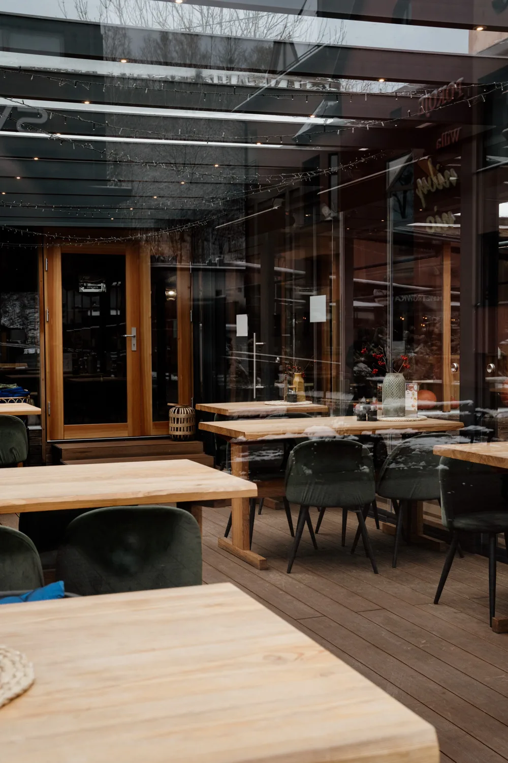 Zadaszony ogródek restauracyjny z drewnianymi stołami i zielonymi krzesłami, widok przez przeszkloną przestrzeń.