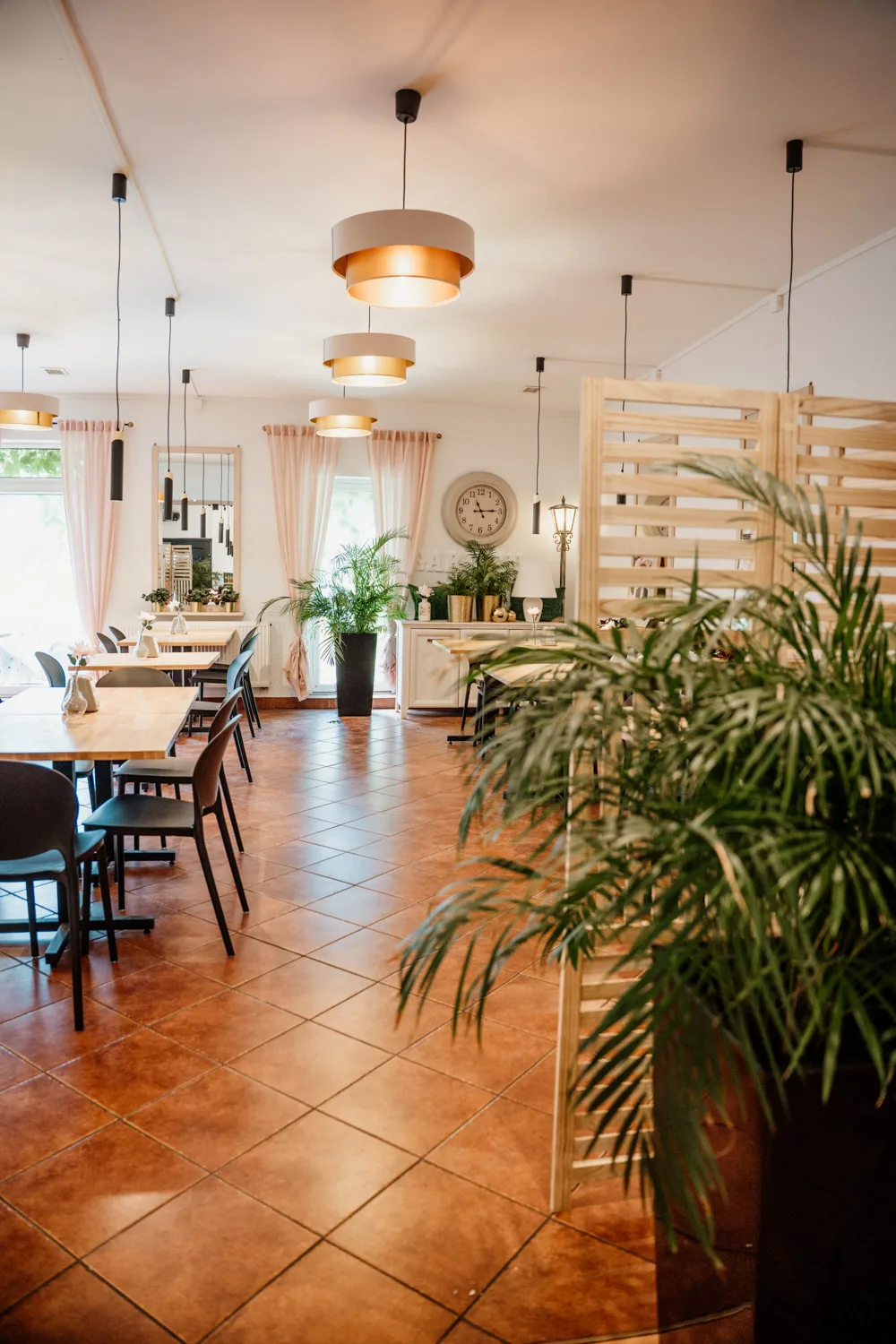 Przytulna sala restauracyjna z jasnymi stolikami, wiszącymi lampami sufitowymi, roślinami doniczkowymi i terakotową podłogą, urządzona w nowoczesnym, ciepłym stylu.