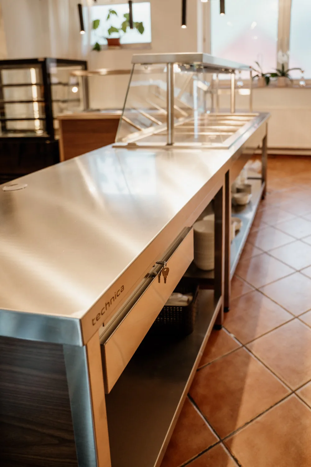 Stalowy stół gastronomiczny z nadstawką i szafką chłodniczą Technica, ustawiony w jasnym zapleczu kuchennym z terakotową podłogą.