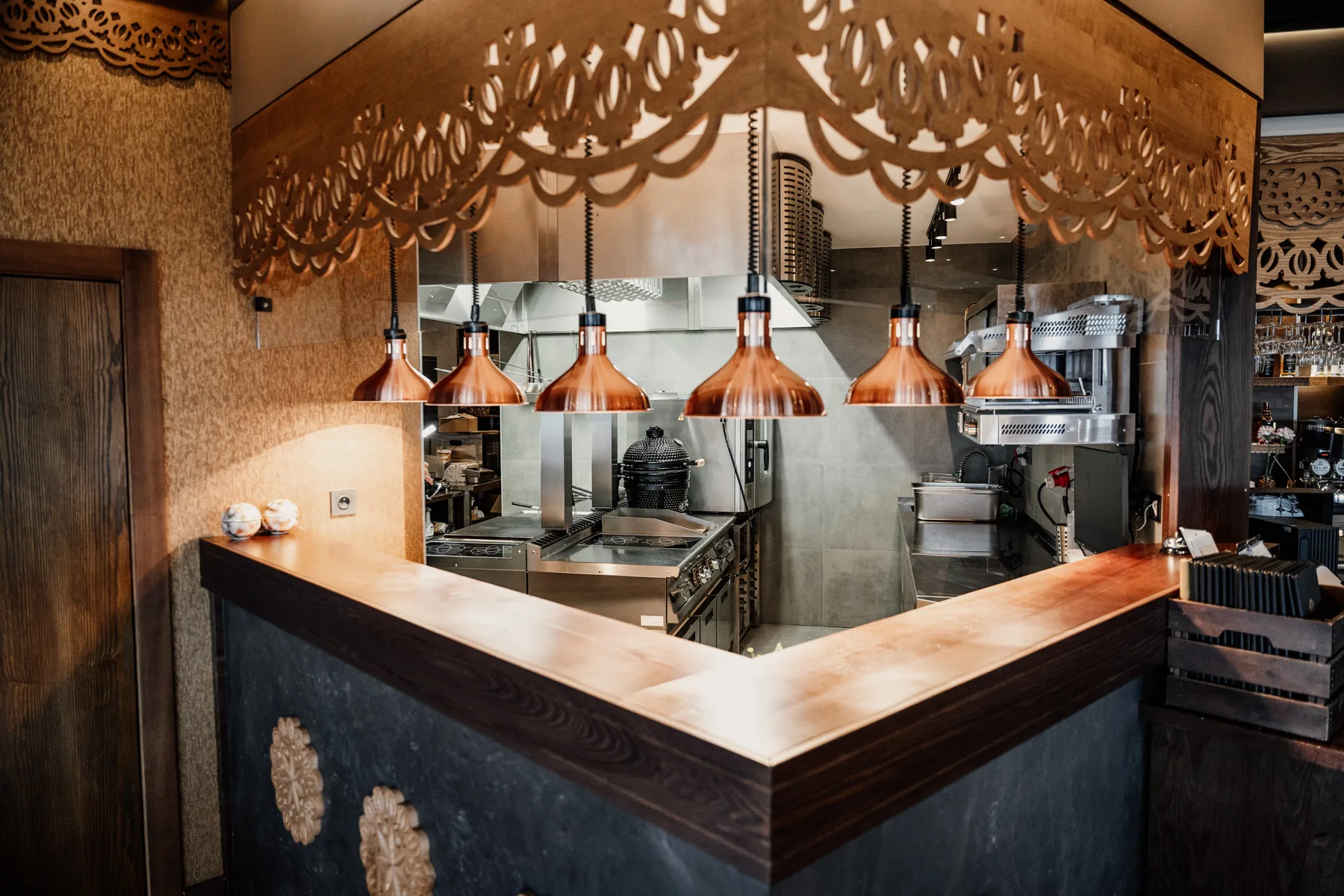 Otwarta kuchnia restauracyjna z dekoracyjną drewnianą zabudową, miedzianymi lampami grzewczymi i profesjonalnym zapleczem gastronomicznym widocznym zza lady.