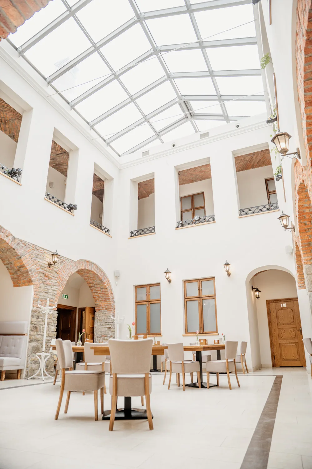 Jasne wnętrze restauracji w historycznym patio z przeszklonym dachem, łukami z cegły i drewnianymi stołami z krzesłami, tworzące elegancką i przestronną salę.