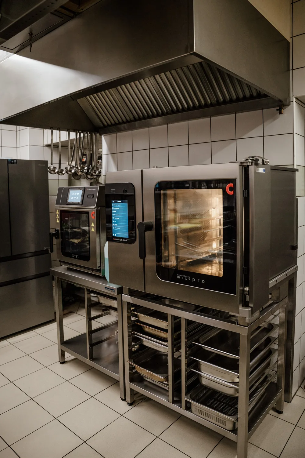 Dwa profesjonalne piece konwekcyjno-parowe w kuchni gastronomicznej, ustawione pod okapem wentylacyjnym, z półkami na pojemniki GN i akcesoria.