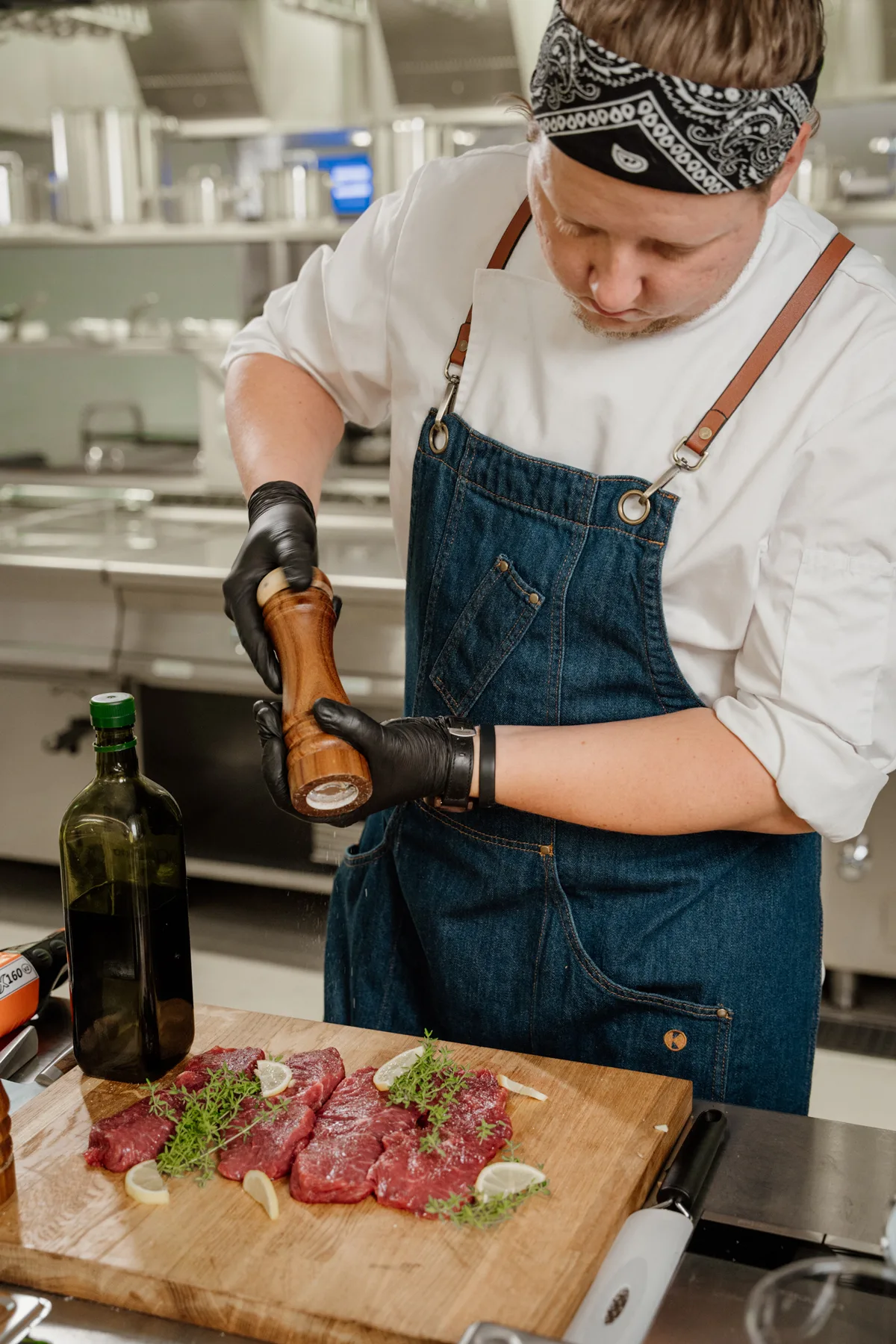 Szef kuchni w profesjonalnej kuchni doprawiający surowe steki solą i pieprzem na drewnianej desce, w otoczeniu sprzętu gastronomicznego.
