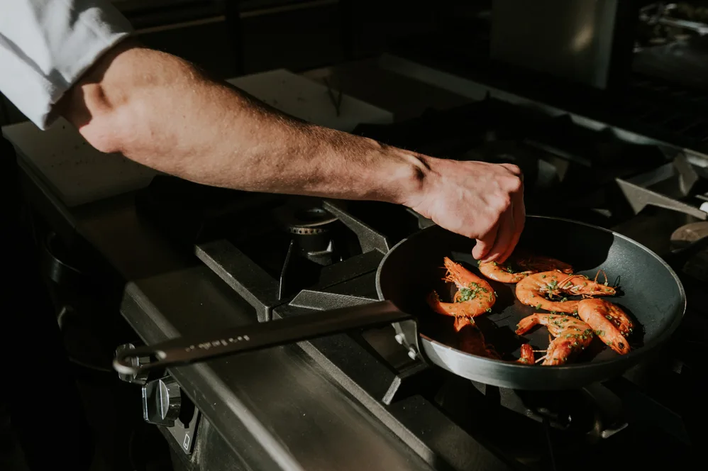 Ręka kucharza smażącego krewetki na patelni na profesjonalnej kuchence gazowej w restauracyjnej kuchni.