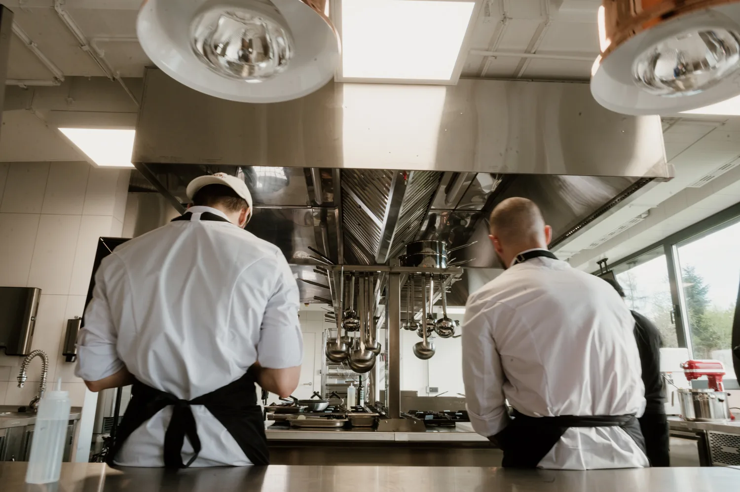 Dwóch kucharzy pracujących przy centralnym stanowisku w profesjonalnej kuchni gastronomicznej z okapem i zawieszonymi chochlami.