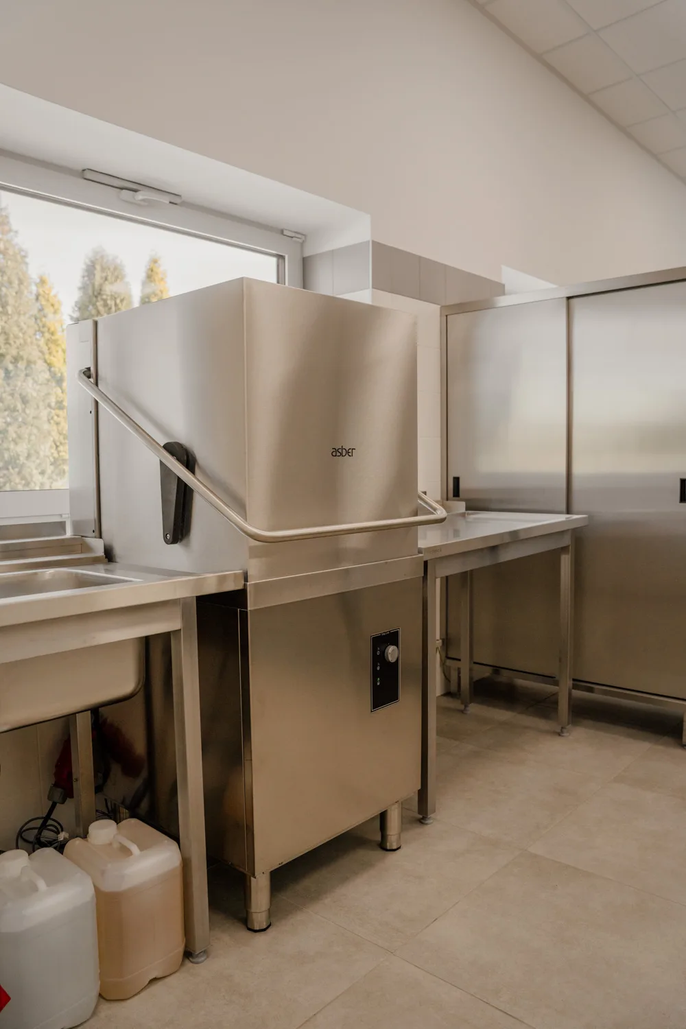 Profesjonalna zmywarka gastronomiczna kapturowa ze stali nierdzewnej, ustawiona w zapleczu kuchennym obok stołów roboczych i zlewu.