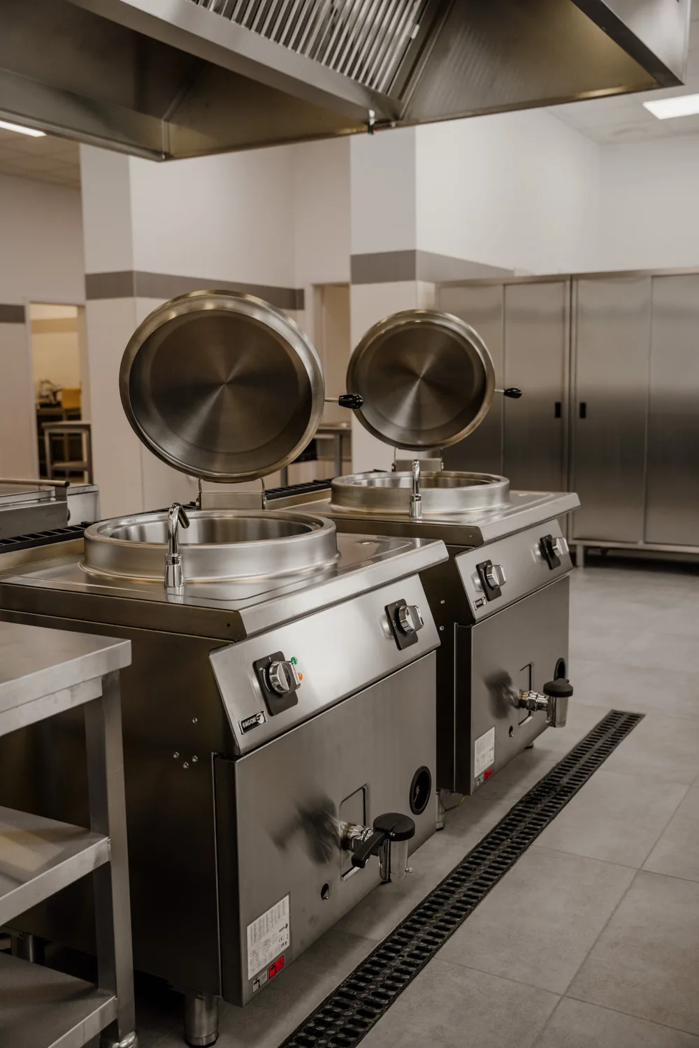 Profesjonalne kotły warzelne ze stali nierdzewnej z podnoszonymi pokrywami, ustawione w nowoczesnej kuchni gastronomicznej pod okapem wentylacyjnym.