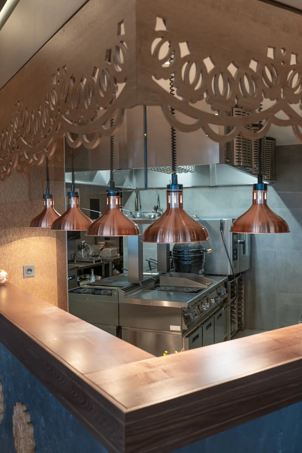 Otwarta kuchnia restauracyjna z dekoracyjną drewnianą zabudową, miedzianymi lampami grzewczymi i profesjonalnym zapleczem gastronomicznym widocznym zza lady.