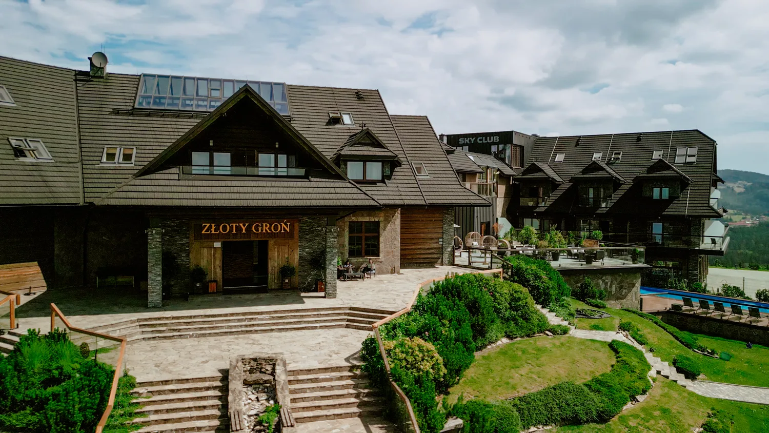 Widok zewnętrzny hotelu i restauracji Złoty Groń w górskim otoczeniu, z tarasami, zielenią i nowoczesną zabudową w tle.