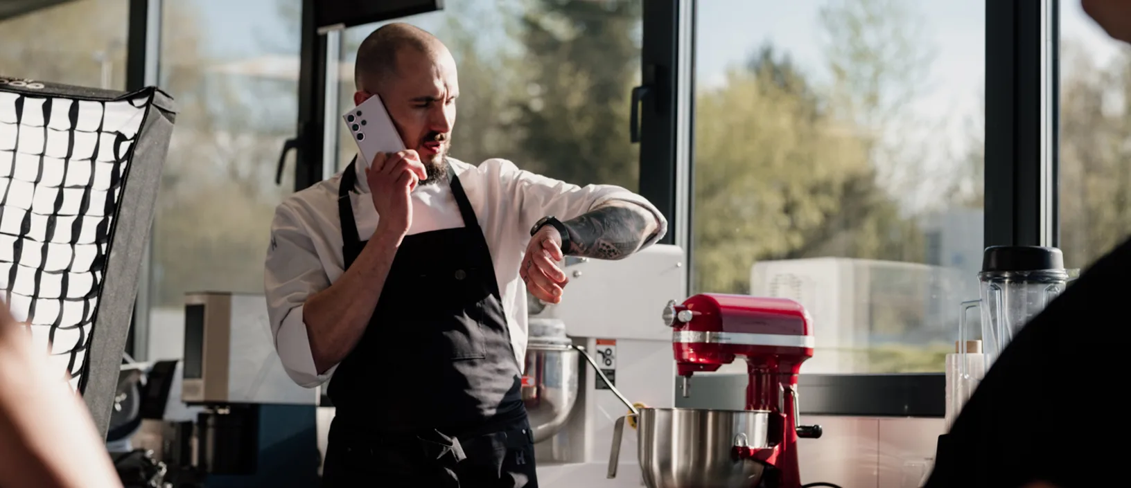 Kucharz w profesjonalnej kuchni rozmawia przez telefon i sprawdza godzinę na zegarku, stojąc przy czerwonym mikserze planetarnym i sprzęcie gastronomicznym.