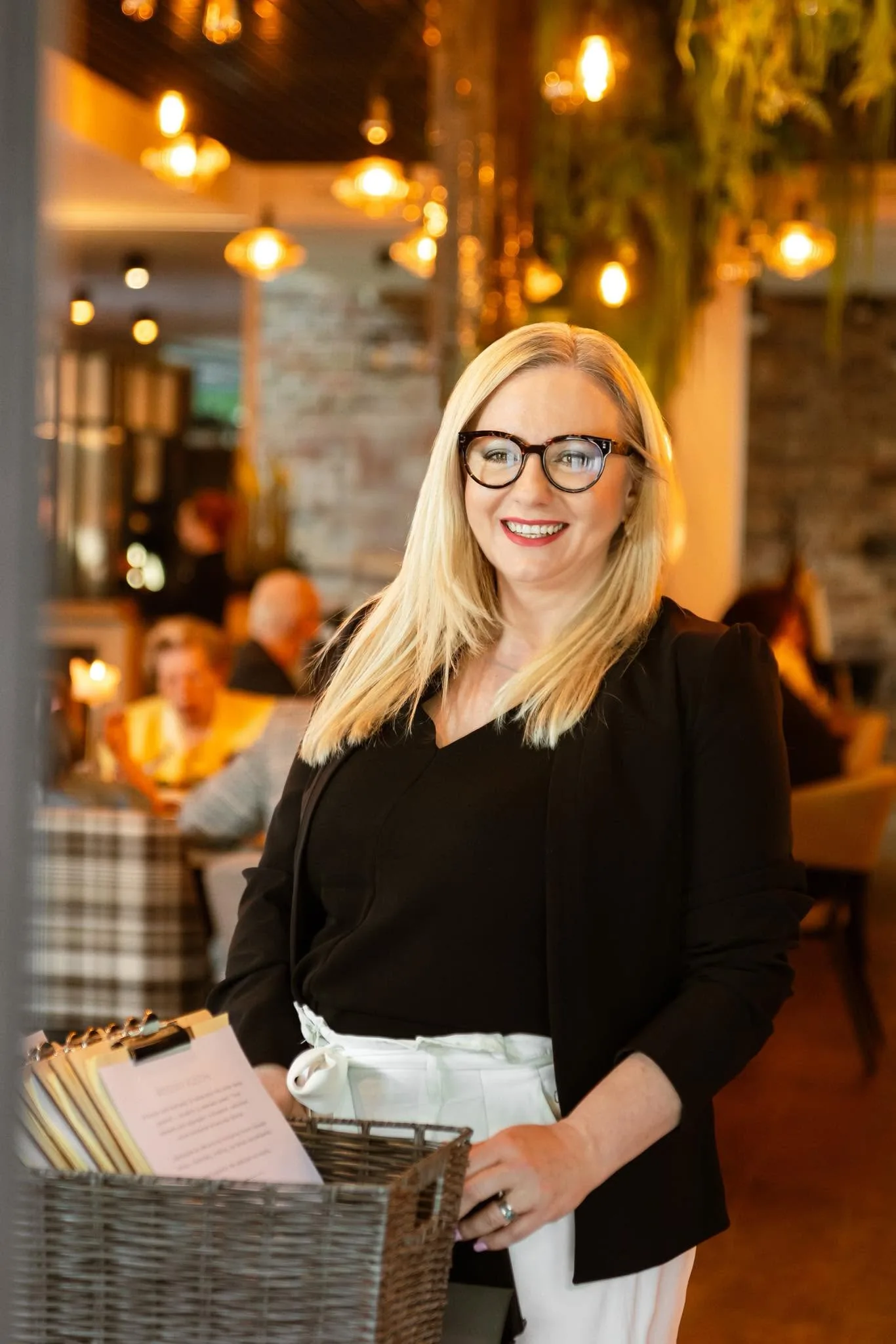 Uśmiechnięta pracownica restauracji stojąca przy wejściu, trzymająca koszyk z kartami menu, na tle ciepło oświetlonego wnętrza z gośćmi przy stolikach.