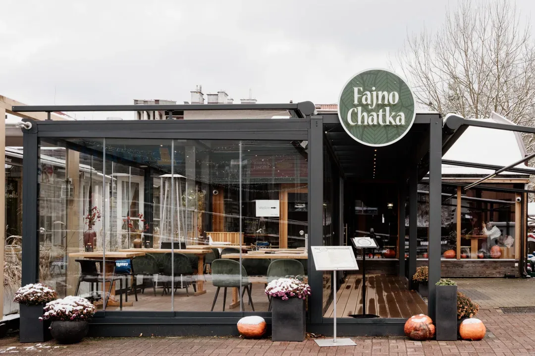 Przeszklony ogródek restauracyjny z widocznym szyldem „Fajno Chatka”, drewnianymi stolikami wewnątrz oraz donicami z roślinami i dekoracyjnymi dyniami przed wejściem.