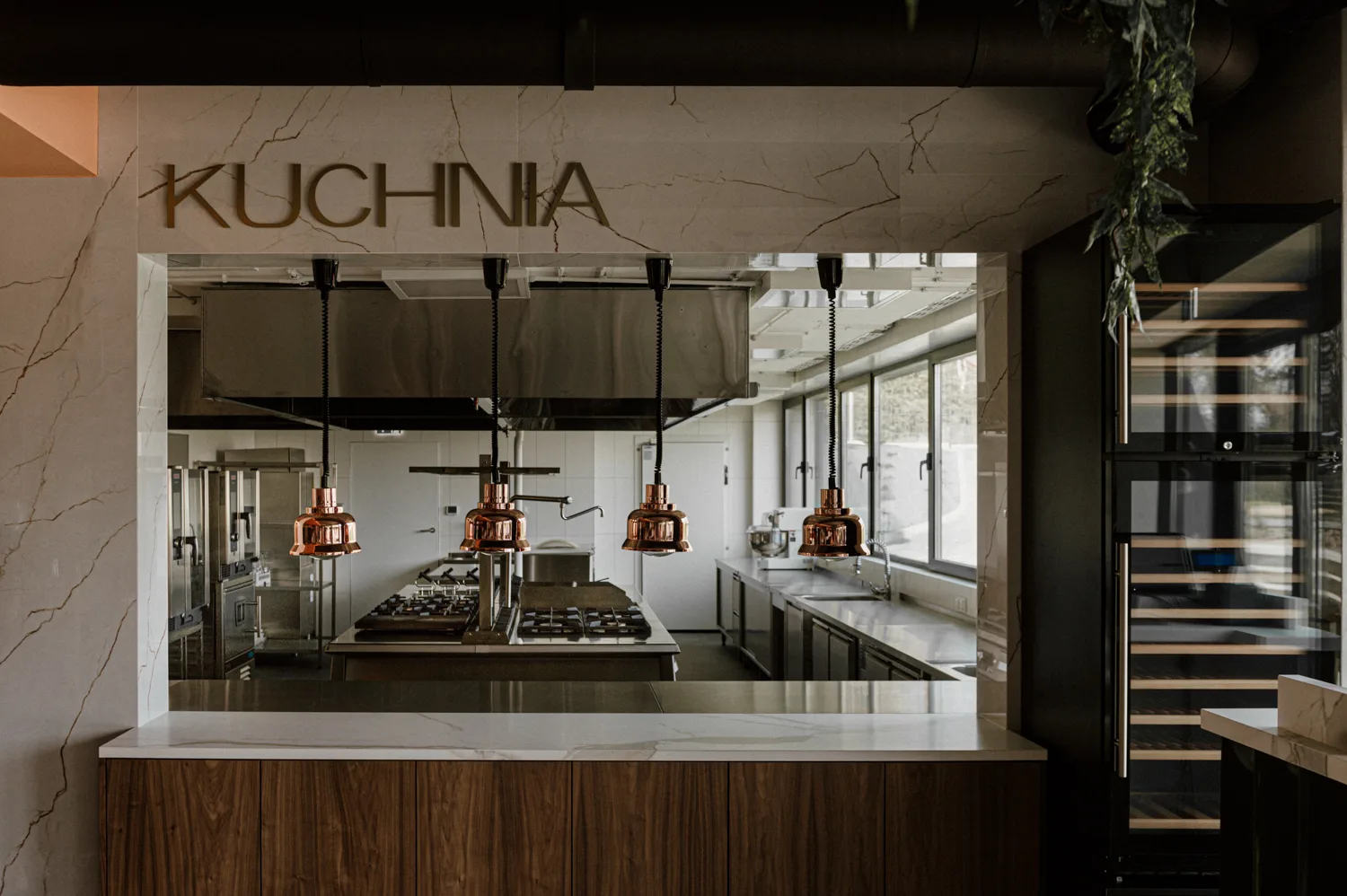 Nowoczesna, otwarta kuchnia restauracyjna z widokiem na profesjonalne stanowiska gotowania, miedziane lampy grzewcze i zabudowę ze stali nierdzewnej.