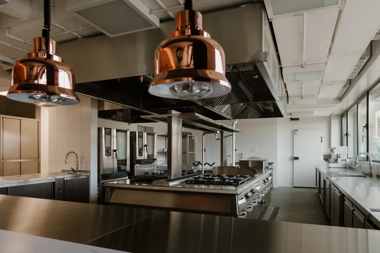 Nowoczesna, otwarta kuchnia restauracyjna z widokiem na profesjonalne stanowiska gotowania, miedziane lampy grzewcze i zabudowę ze stali nierdzewnej.