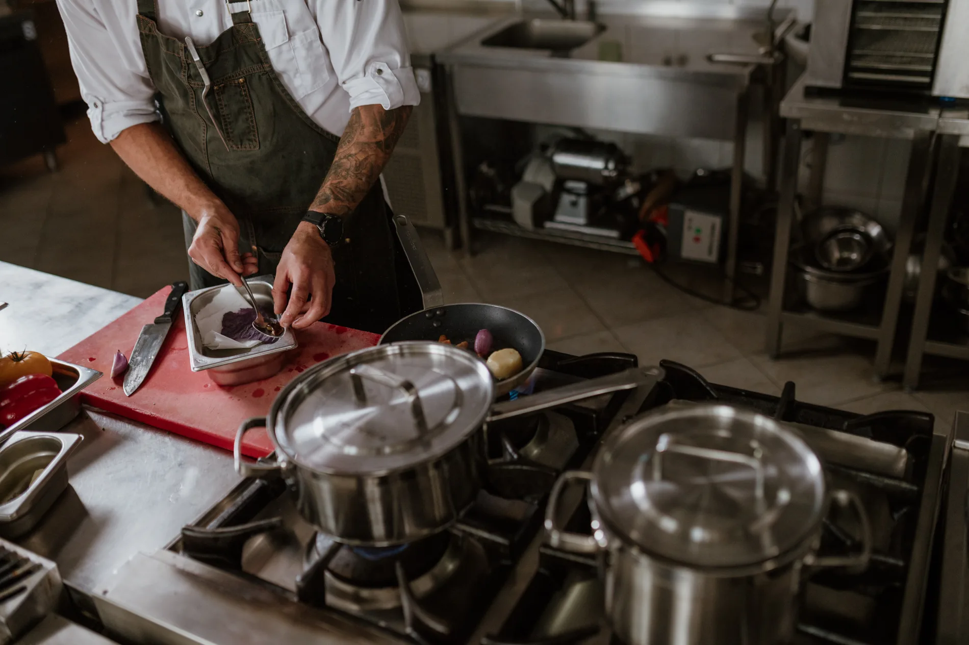 Zbliżenie na szefa kuchni, z patelniami na palnikach i zapleczem sprzętu HoReCa w tle.