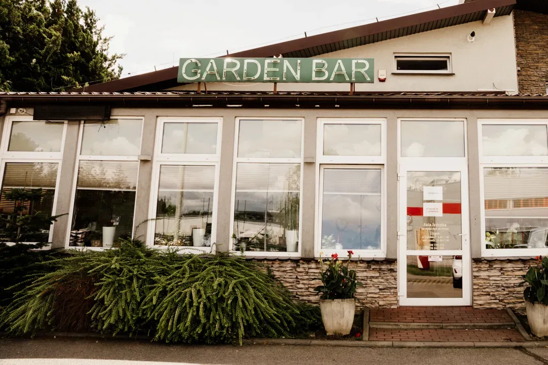 Elewacja restauracji Garden Bar z dużymi oknami, zielonym szyldem nad wejściem oraz roślinami ozdobnymi przed budynkiem.