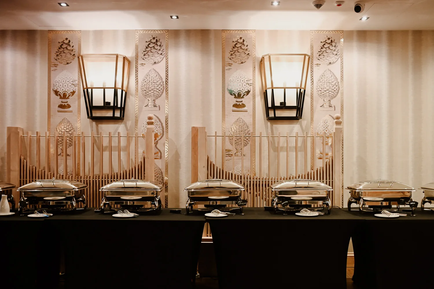 Elegancka strefa bufetowa z podgrzewaczami gastronomicznymi ustawionymi na czarnych stołach, na tle dekoracyjnej ściany z ornamentami i stylowym oświetleniem.