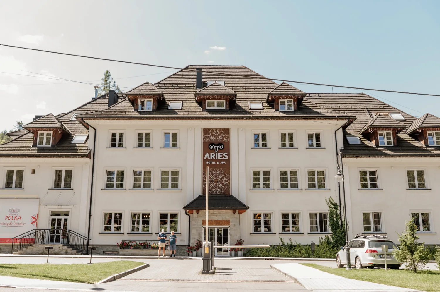 Elewacja hotelu Aries Hotel & Spa – jasny, elegancki budynek z centralnym wejściem, logo Aries na fasadzie oraz gośćmi przed wejściem, w otoczeniu zieleni.