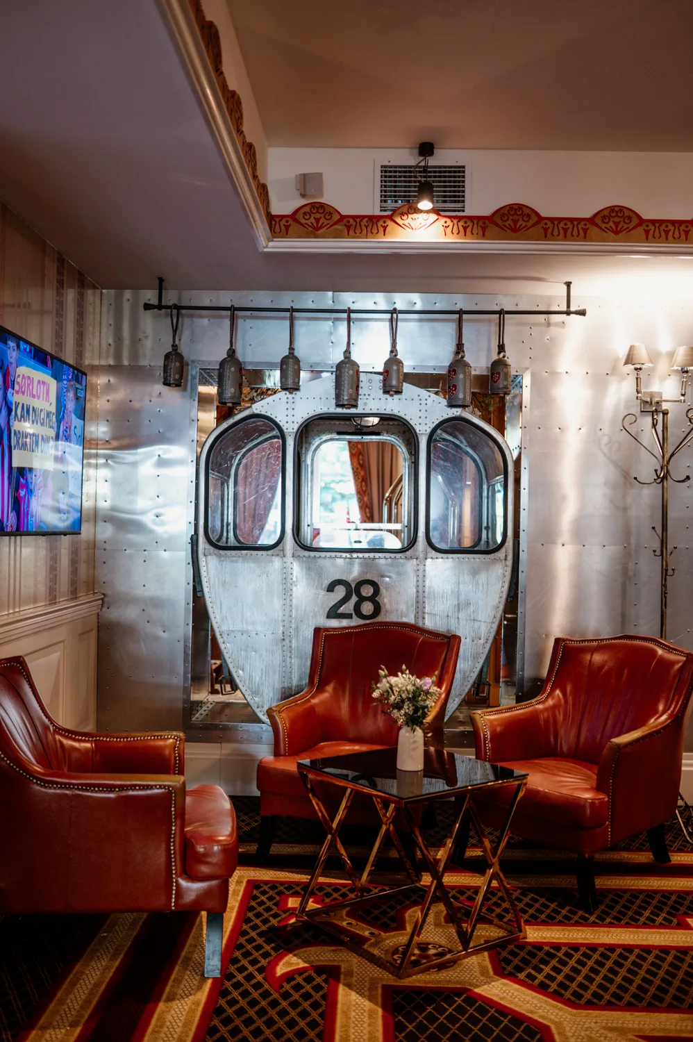 Stylowe wnętrze salonu hotelowego z czerwonymi skórzanymi fotelami i stolikiem kawowym, z industrialnym elementem dekoracyjnym w formie metalowych drzwi oznaczonych numerem 28.