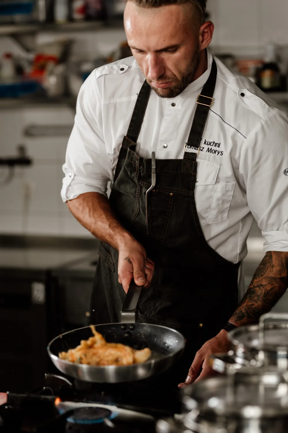 Szef kuchni w profesjonalnej kuchni gastronomicznej, pracujący przy płycie grzewczej z garnkami ze stali nierdzewnej, w trakcie przygotowywania potraw.