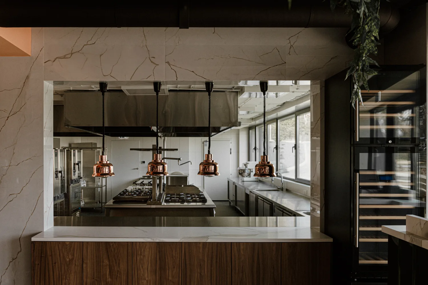 Nowoczesna, otwarta kuchnia restauracyjna z widokiem na profesjonalne stanowiska gotowania, miedziane lampy grzewcze i zabudowę ze stali nierdzewnej.
