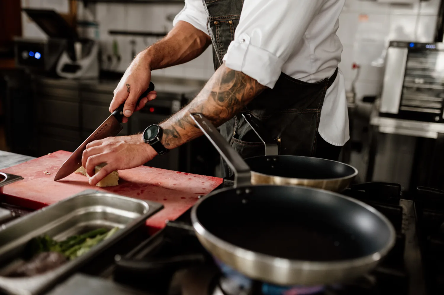 Zbliżenie na szefa kuchni krojącego składniki nożem na desce w profesjonalnej kuchni gastronomicznej, z patelniami na palnikach i zapleczem sprzętu HoReCa w tle.