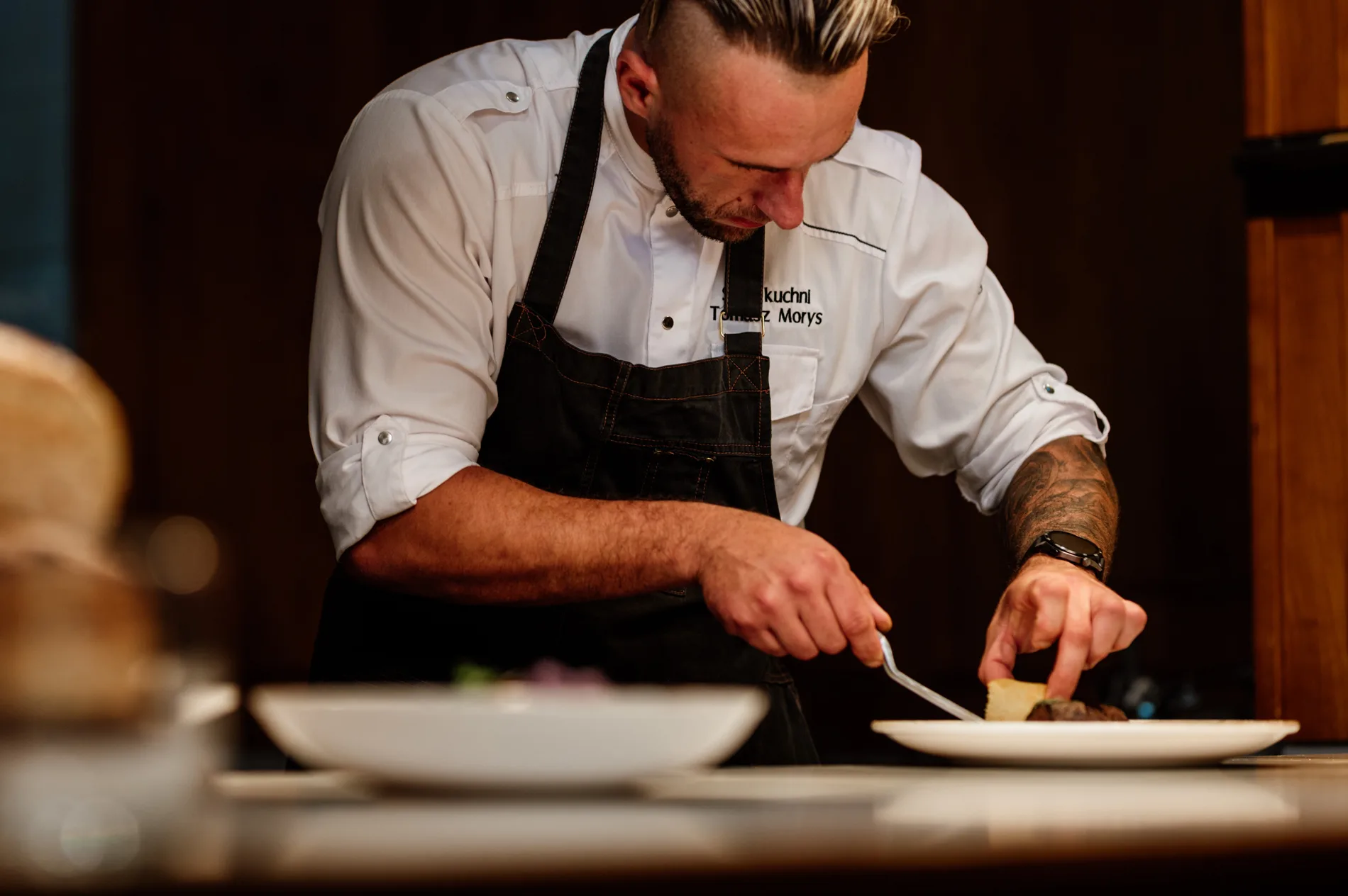 Szef kuchni w białej kurtce i fartuchu precyzyjnie nakłada składniki na talerz, wykańczając elegancką potrawę w restauracyjnej kuchni.