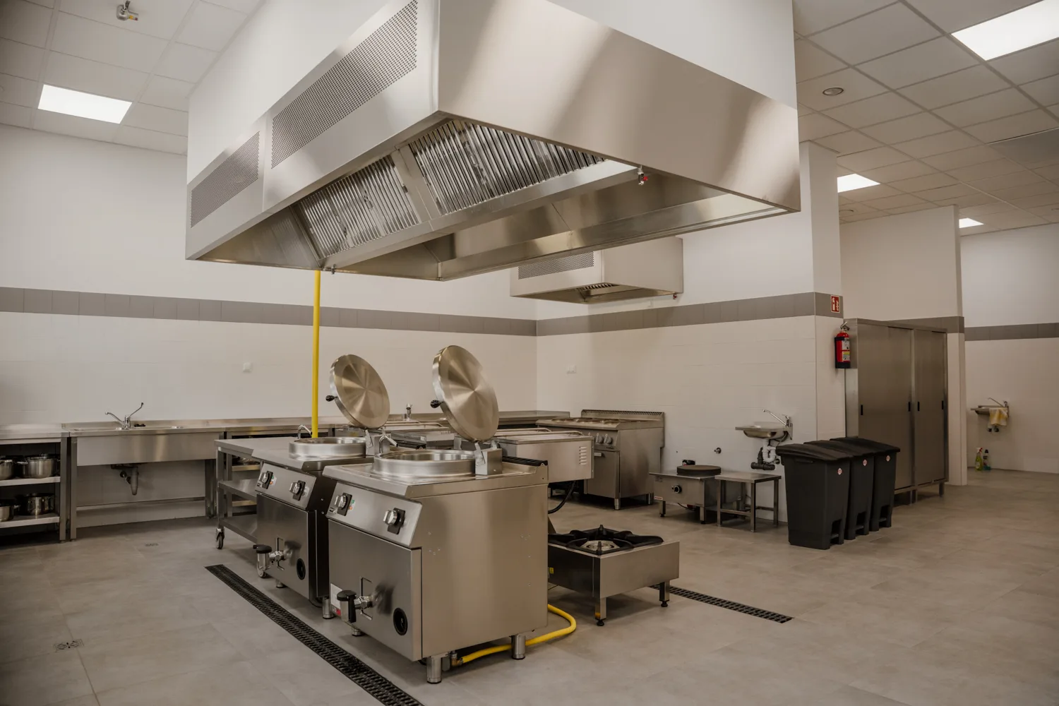Profesjonalna kuchnia gastronomiczna z centralnie ustawionymi urządzeniami grzewczymi i dużymi okapami wentylacyjnymi ze stali nierdzewnej, przygotowana do intensywnej pracy w obiekcie HoReCa.