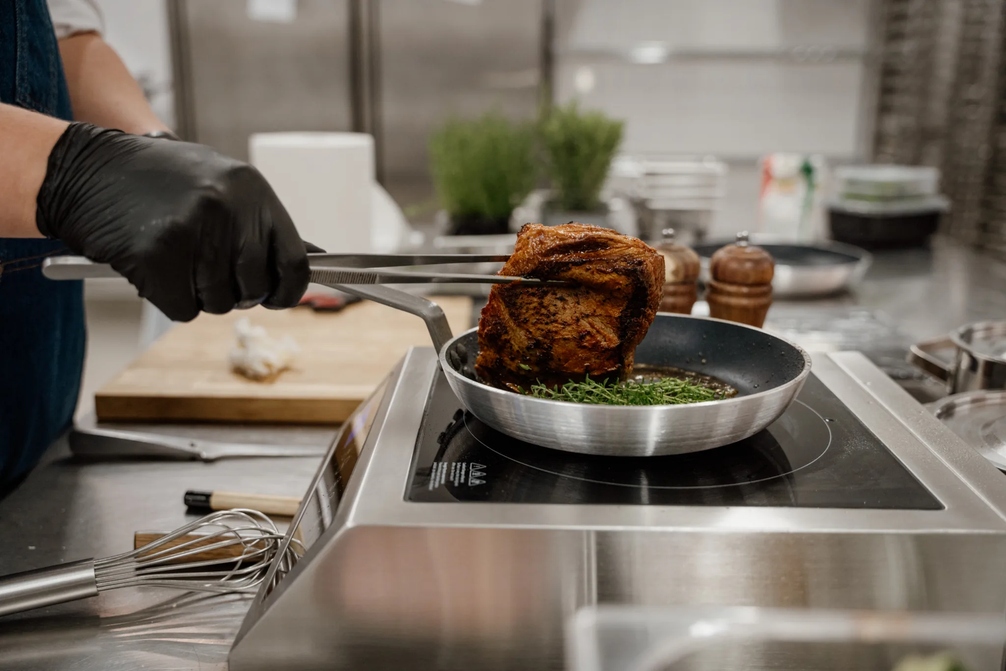 Smażenie mięsa w profesjonalnej kuchni gastronomicznej – szef kuchni obraca stek na patelni na płycie indukcyjnej.