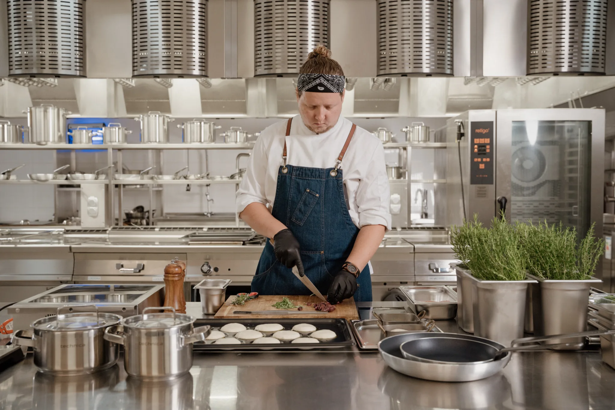 Szef kuchni pracujący w profesjonalnej kuchni gastronomicznej – krojenie składników na blacie ze stali nierdzewnej, w otoczeniu sprzętu i naczyń gastronomicznych.