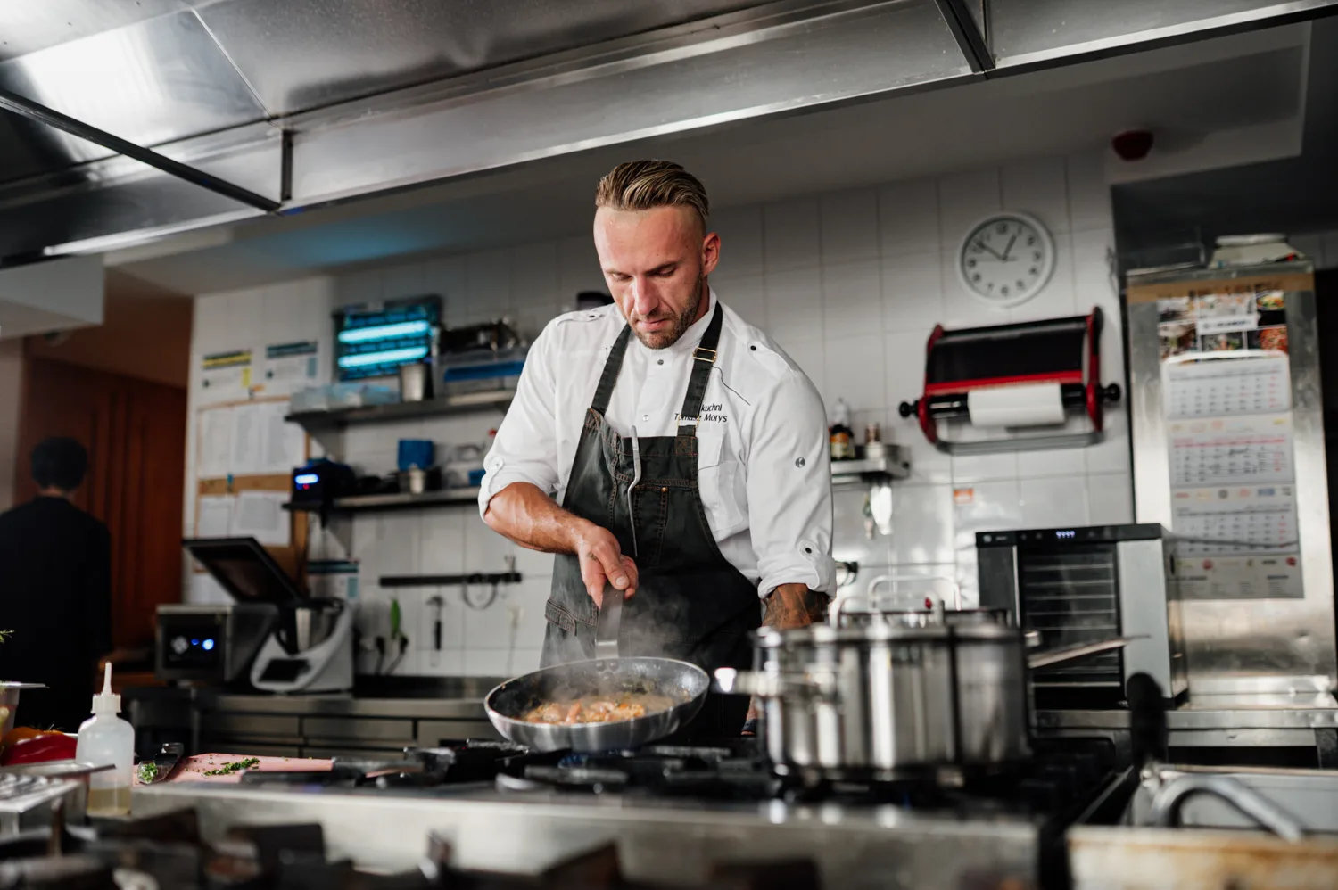 Szef kuchni podczas pracy w profesjonalnej kuchni gastronomicznej, przygotowujący danie na patelni przy stanowisku z nowoczesnym sprzętem kuchennym.