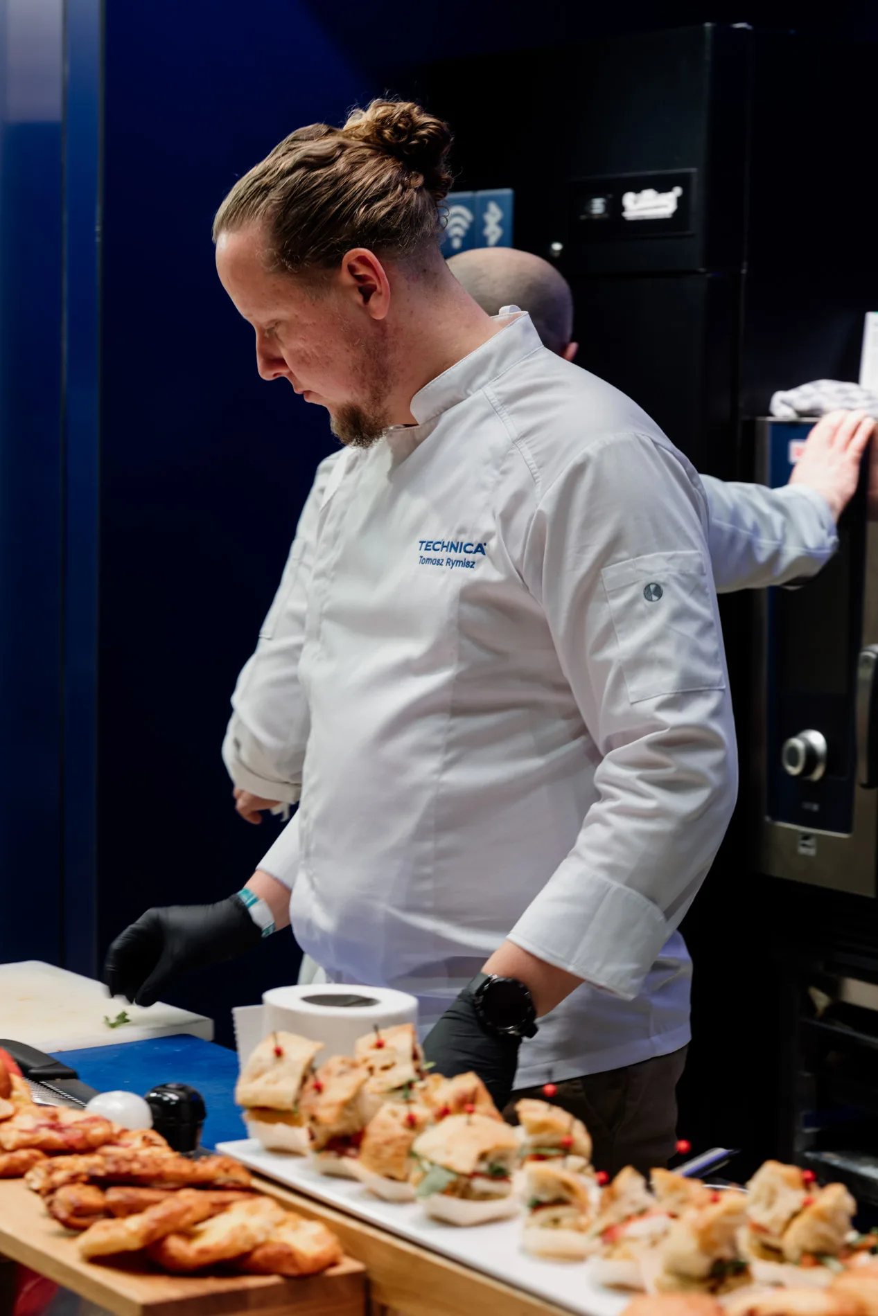 Szef kuchni w fartuchu z logo Technica przygotowujący przekąski podczas pokazu targów przy profesjonalnym sprzęcie gastronomicznym.