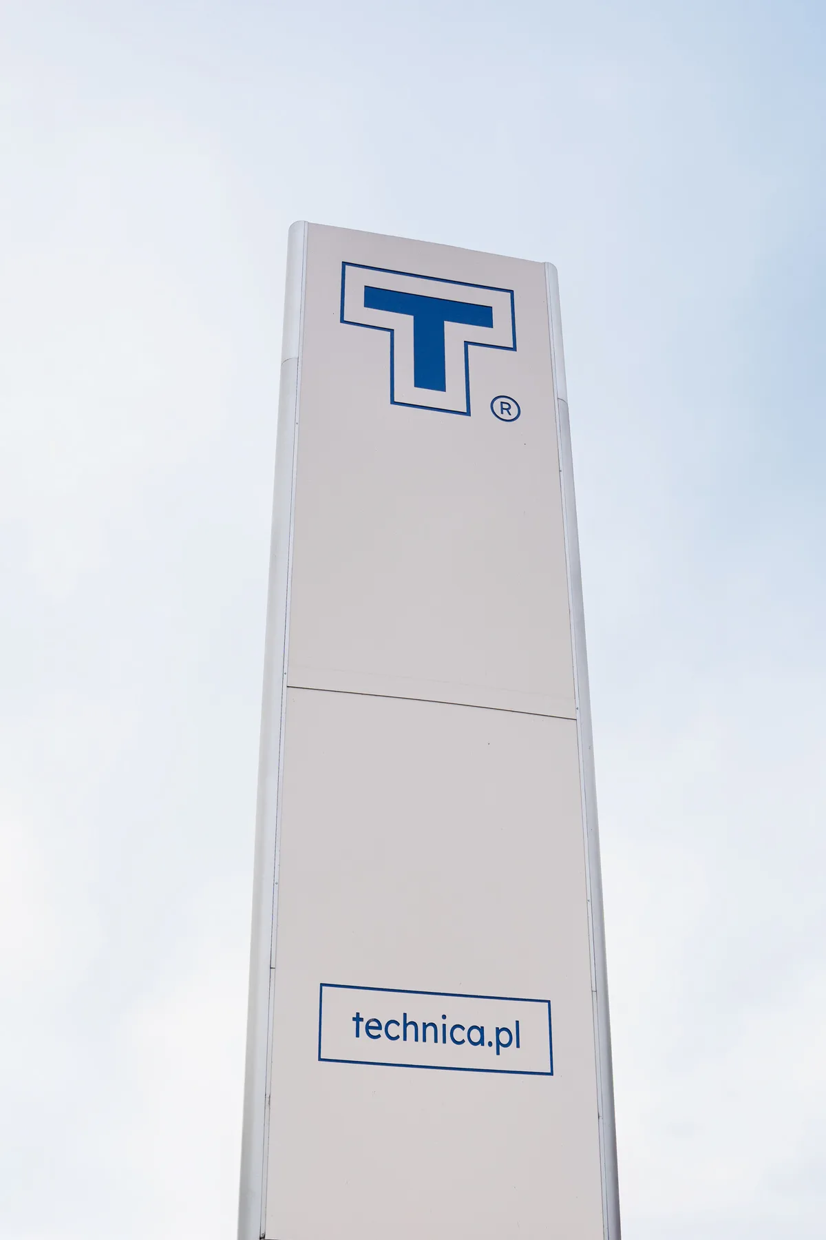 Pylon reklamowy marki Technica z logo „T” oraz adresem technica.pl na jasnym tle nieba.